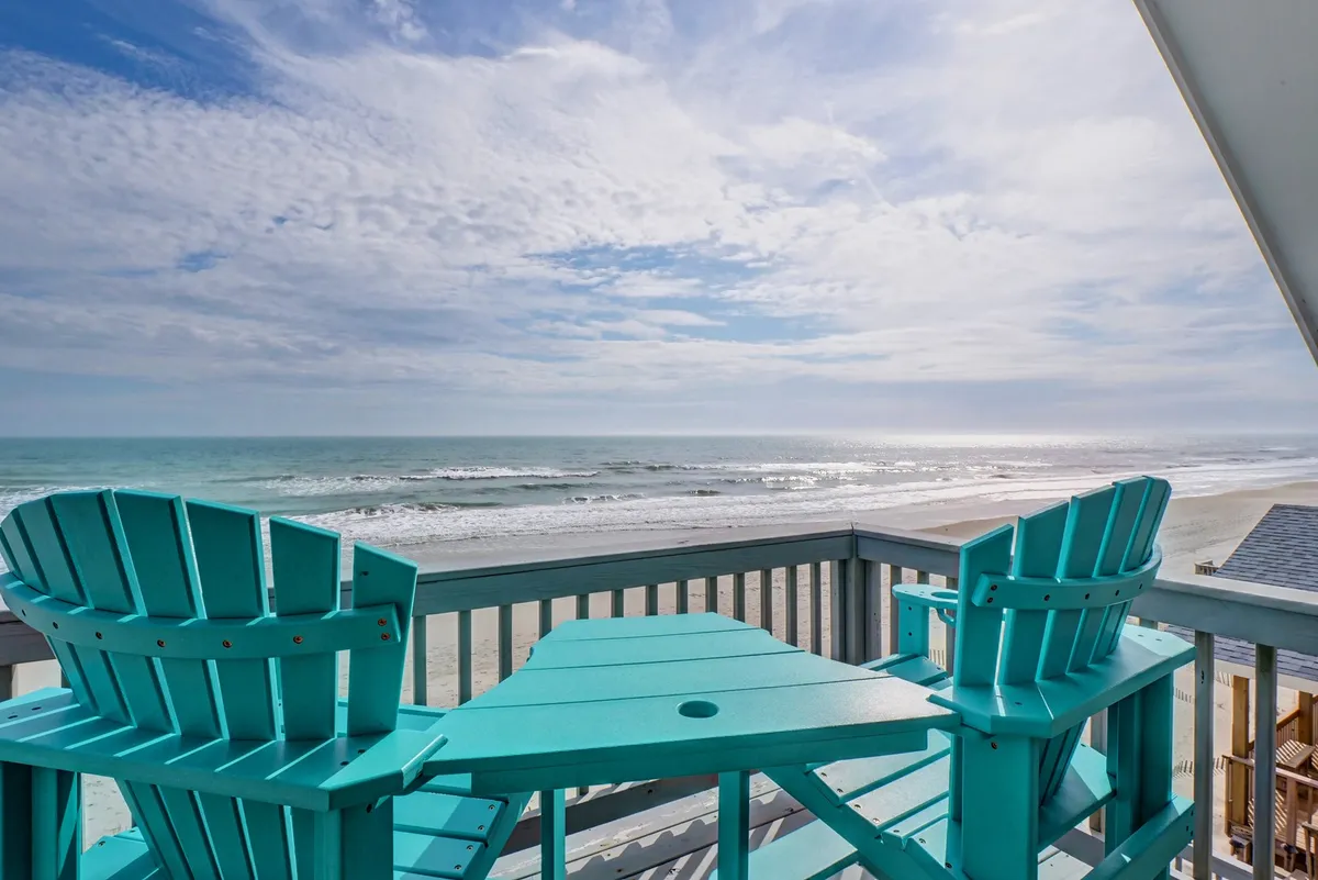 A pair of turquoise chairs are arranged on a wooden deck overlooking the ocean and framed by a clear blue sky. The scene is tranquil, inviting relaxation, with the ocean stretching beyond the railing—ideal for sunrise coffee or sunset toasts.