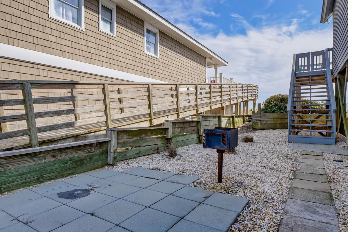 An exterior shot focused on a wooden ramp and steps leading up to the house, a grill area, and a pebbled ground for outdoor gatherings. The relaxed, practical vibe suggests accessibility and amenities for travelers arriving by car.
