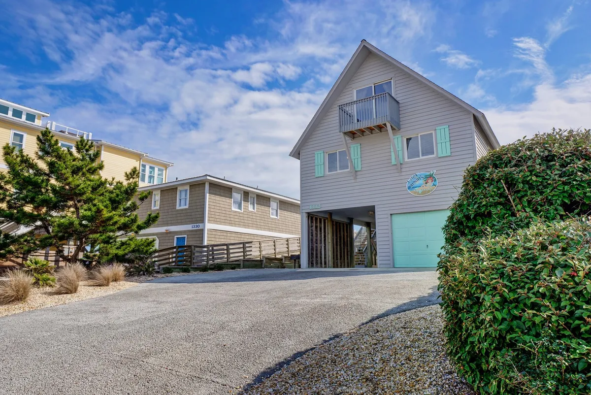 A cheerful exterior shot of the house facade, driveway, and balcony with vibrant green shutters. The driveway leads up to the front, visually reinforcing ease of arrival and private entry.