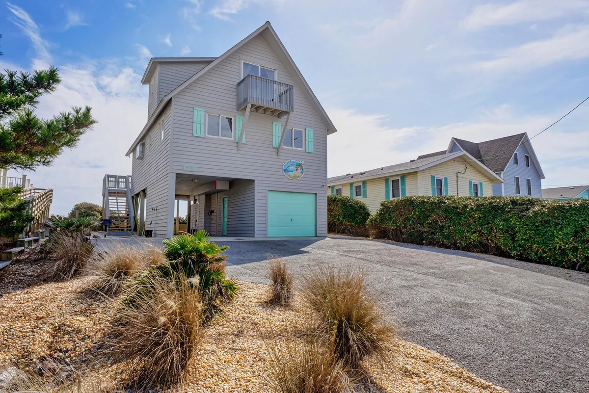 This image shows the exterior of a beach house with a balcony, surrounded by lush greenery and featuring a decorative sign on the house. The scene exudes a sense of arrival, outdoor relaxation, and the beach environment that the property is situated in.