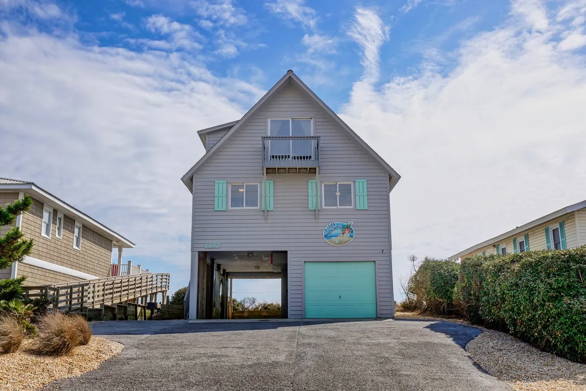 This exterior image highlights the house’s unique architectural design with a vibrant turquoise garage door and a decorative fish sign. The driveway and garage area are clearly visible, emphasizing the carport and entry point for guests.
