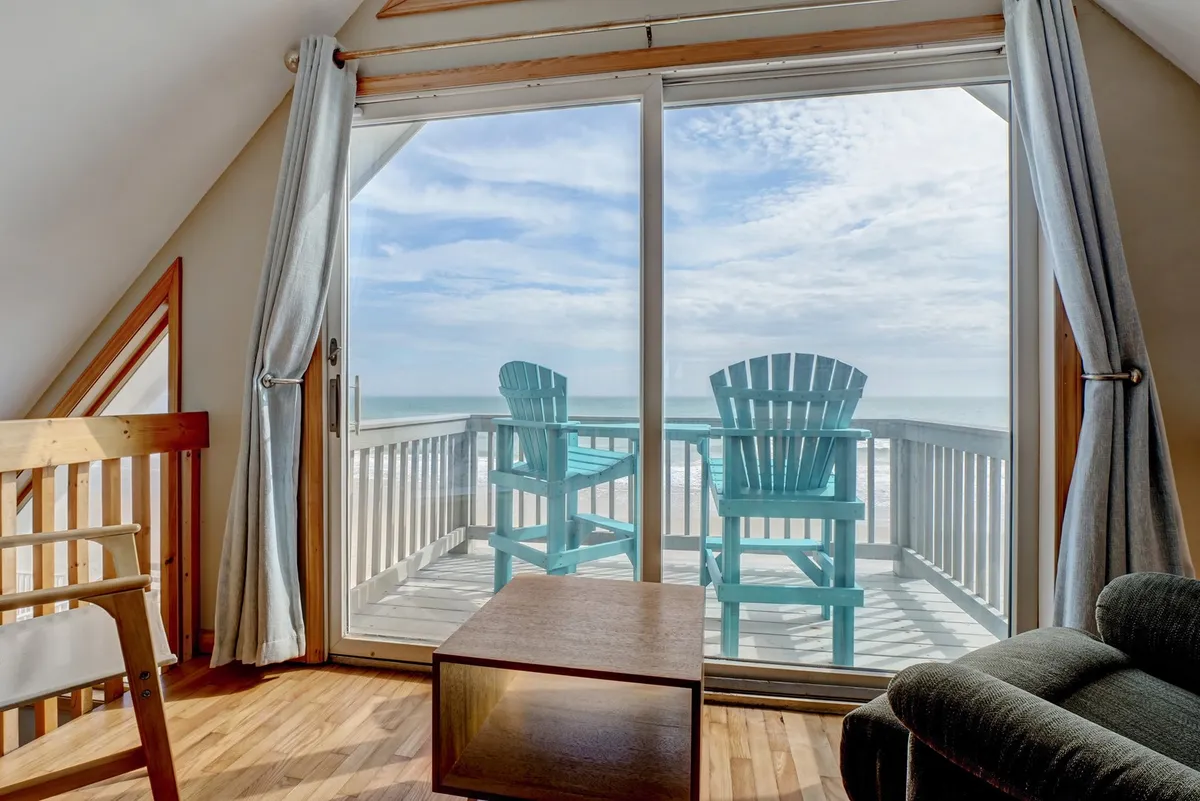 This image features a spacious living area with a large sliding glass door opening to a deck furnished with teal Adirondack chairs. The arrangement allows for seamless indoor-outdoor living, with comfortable seating positioned to enjoy ocean views and natural light filling the room.