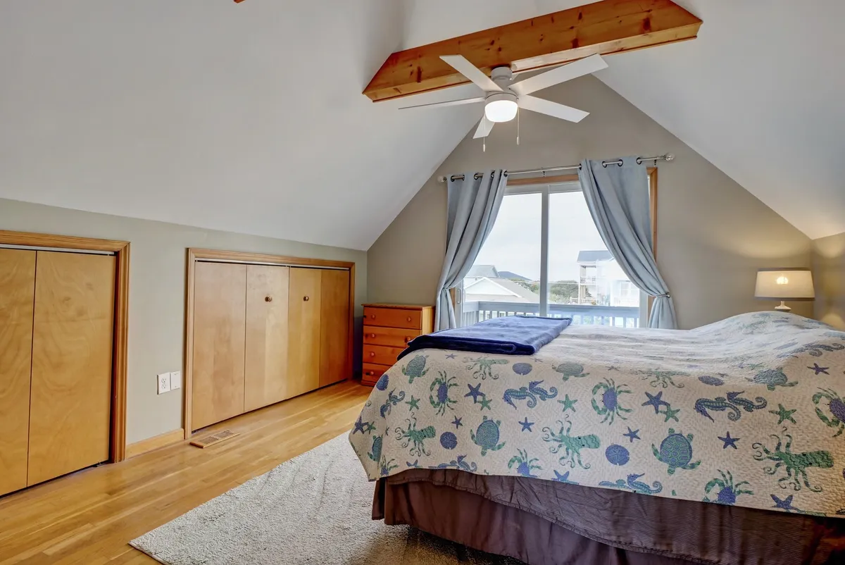 A tranquil upstairs bedroom with an ocean-themed quilt, large window offering a view, and wooden closet doors. The natural light creates a peaceful, serene environment, perfect for relaxation, morning coffee, or listening to distant waves.