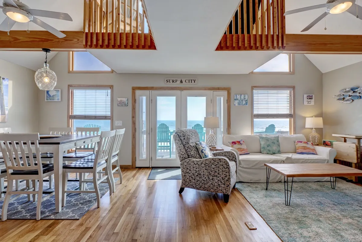 A spacious interior with a large dining table, a comfortable seating area, and expansive windows that frame a stunning ocean view. Decorative touches, such as a Surf City sign and coastal artwork, add to the character-filled ambience.