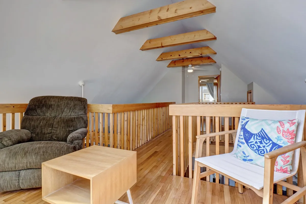 An airy loft area with a comfortable recliner, wooden railings, exposed ceiling beams, and a side table. The space is inviting for reading, conversation, or enjoying elevated views of the main living area. The design emphasizes openness and relaxation.