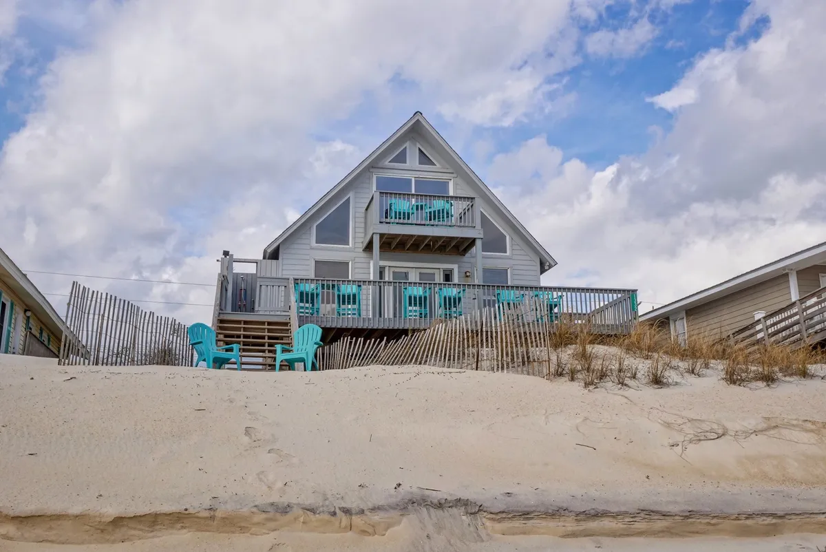 A relaxed beach house scene with turquoise chairs arranged on a deck, a sturdy railing, and the beach just steps away. The mood is casual and serene, perfect for lounging with an ocean backdrop.
