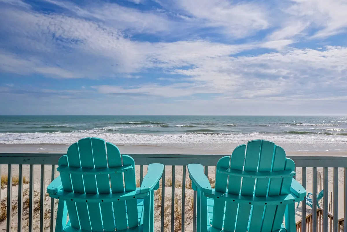A pair of turquoise Adirondack chairs sit on a deck overlooking gentle ocean waves under a clear blue sky. The view is open and serene, ideal for quiet reflection or sharing a romantic sunrise together.