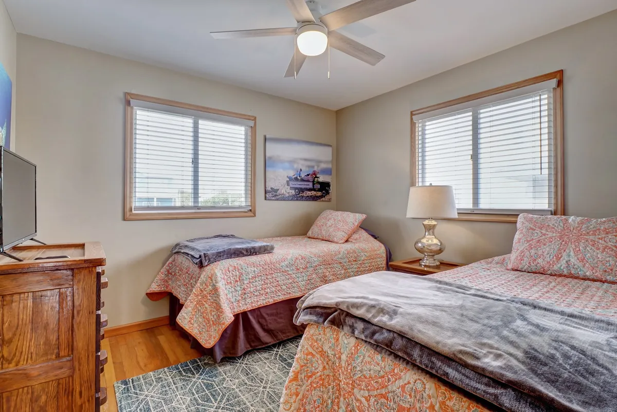 A bedroom with two beds, each adorned with colorful patterned quilts. A nightstand with a lamp sits between the beds, and the walls are decorated with local fishing-themed art. The room is bathed in natural light, suggesting a peaceful space for families or friends to unwind together.