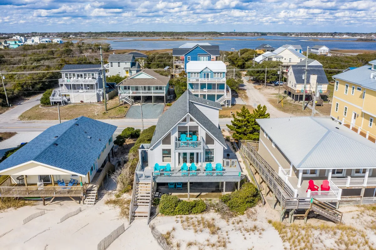 A cheerful exterior shot of a beachfront house, vibrant chairs on the deck overlooking the ocean, and neighboring houses in view. The scene suggests spaces to gather, relax, and enjoy ocean vistas, with the deck positioned as a hub for family gatherings between beach outings.