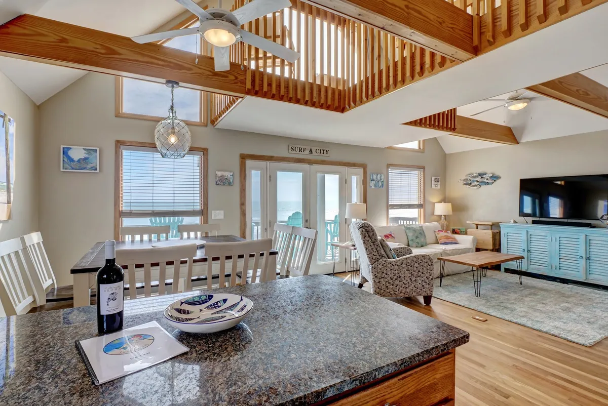 This image showcases an open living and dining area with a granite countertop, dining table and chairs, a comfortable seating area, large windows with direct ocean views, and decorative art on the walls. The bright, airy atmosphere is accentuated by natural light streaming in, inviting guests to relax and connect.