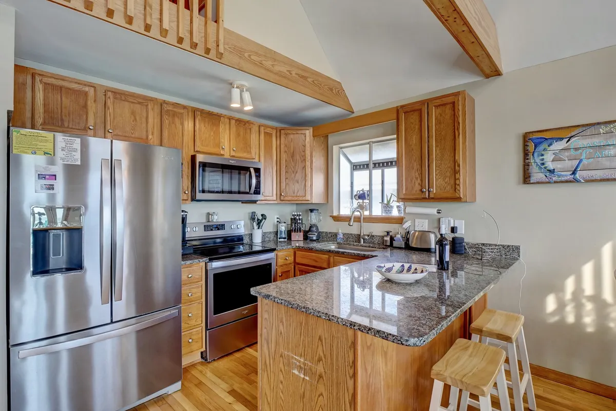 A modern, fully equipped kitchen with stainless steel appliances, granite countertops, wooden cabinets, a central island, and a window letting in natural light. The layout is functional and welcoming, ideal for preparing meals and gathering as a family.