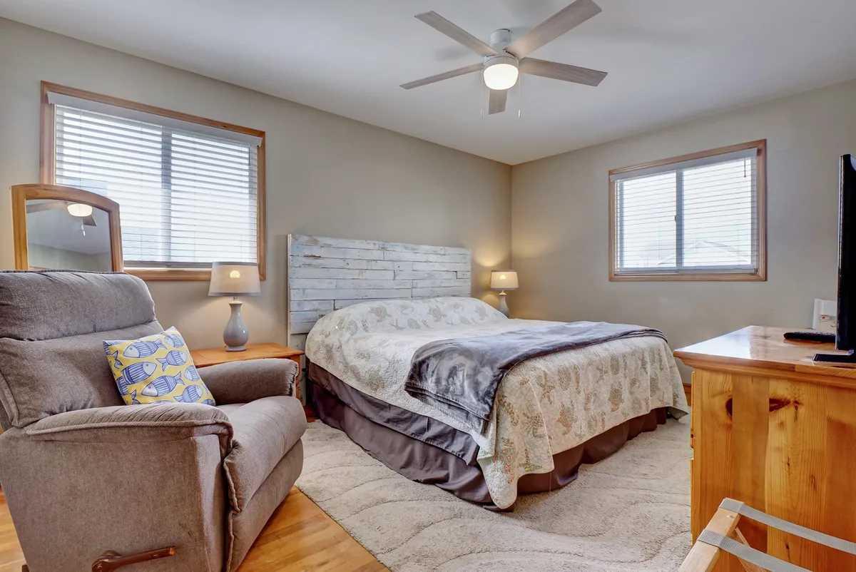 A cozy bedroom featuring a comfortable bed with soft linens, an armchair positioned near the window, nightstands with warm lighting, and a TV mounted across from the bed. Sunlight streams in, creating a welcoming and peaceful ambiance ideal for morning relaxation or a quiet evening.