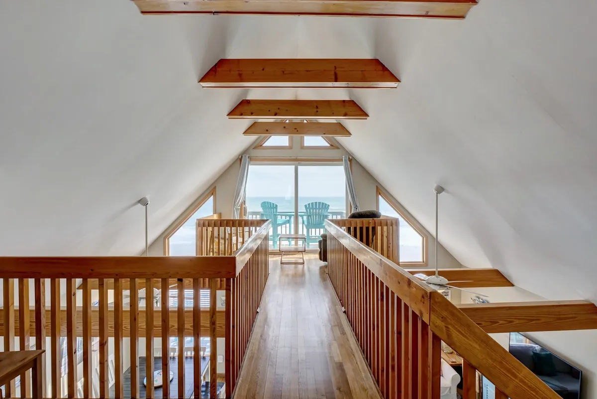 A tranquil interior featuring large windows with breathtaking ocean views, exposed wooden beams in the ceiling, and Adirondack chairs on the balcony. The space is bathed in natural light and designed for relaxation, with a seamless connection to the outdoors.